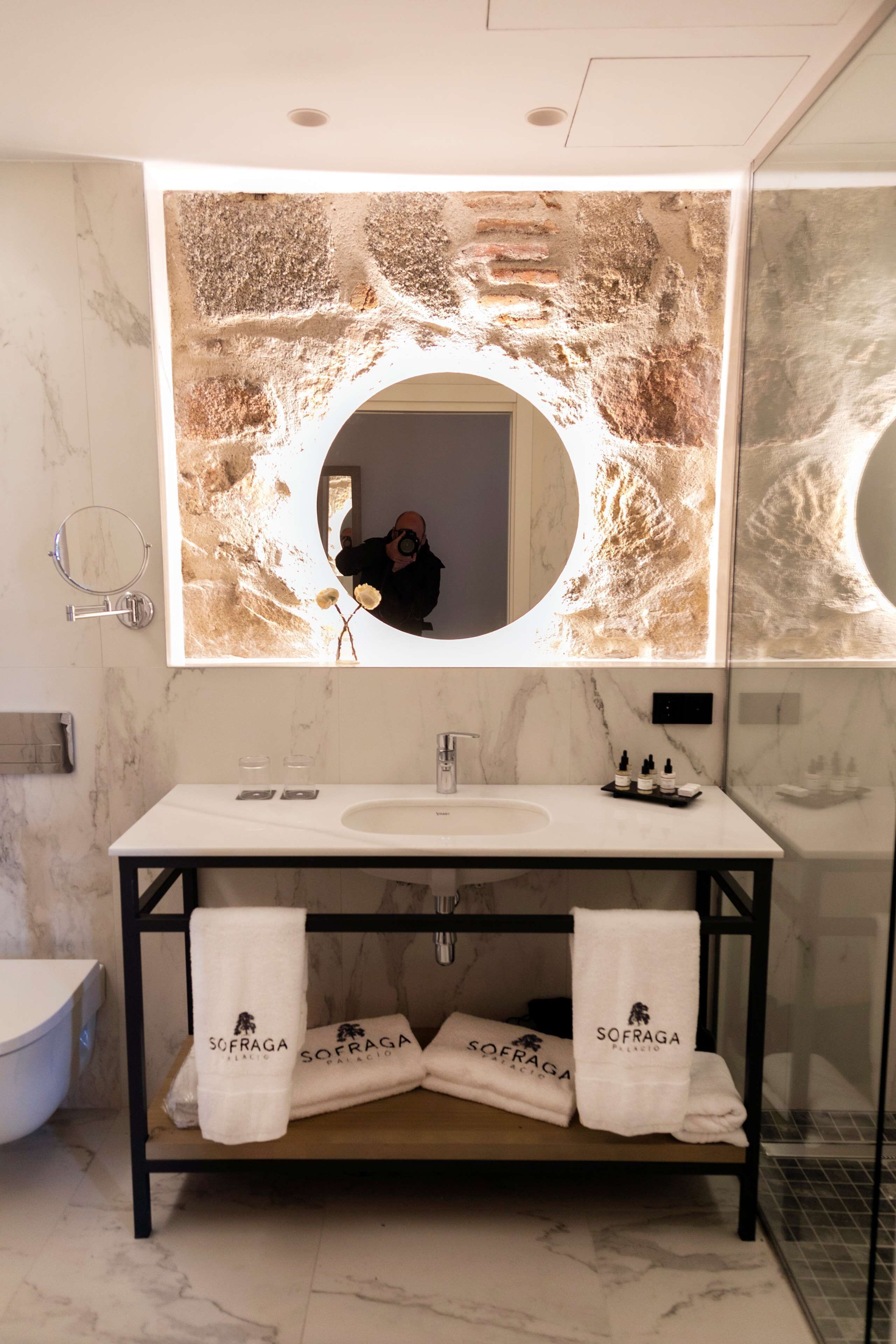 A modern bathroom with a marble sink, a large mirror with a circular frame, and two towels with the brand "SOFRAGA" on a shelf.