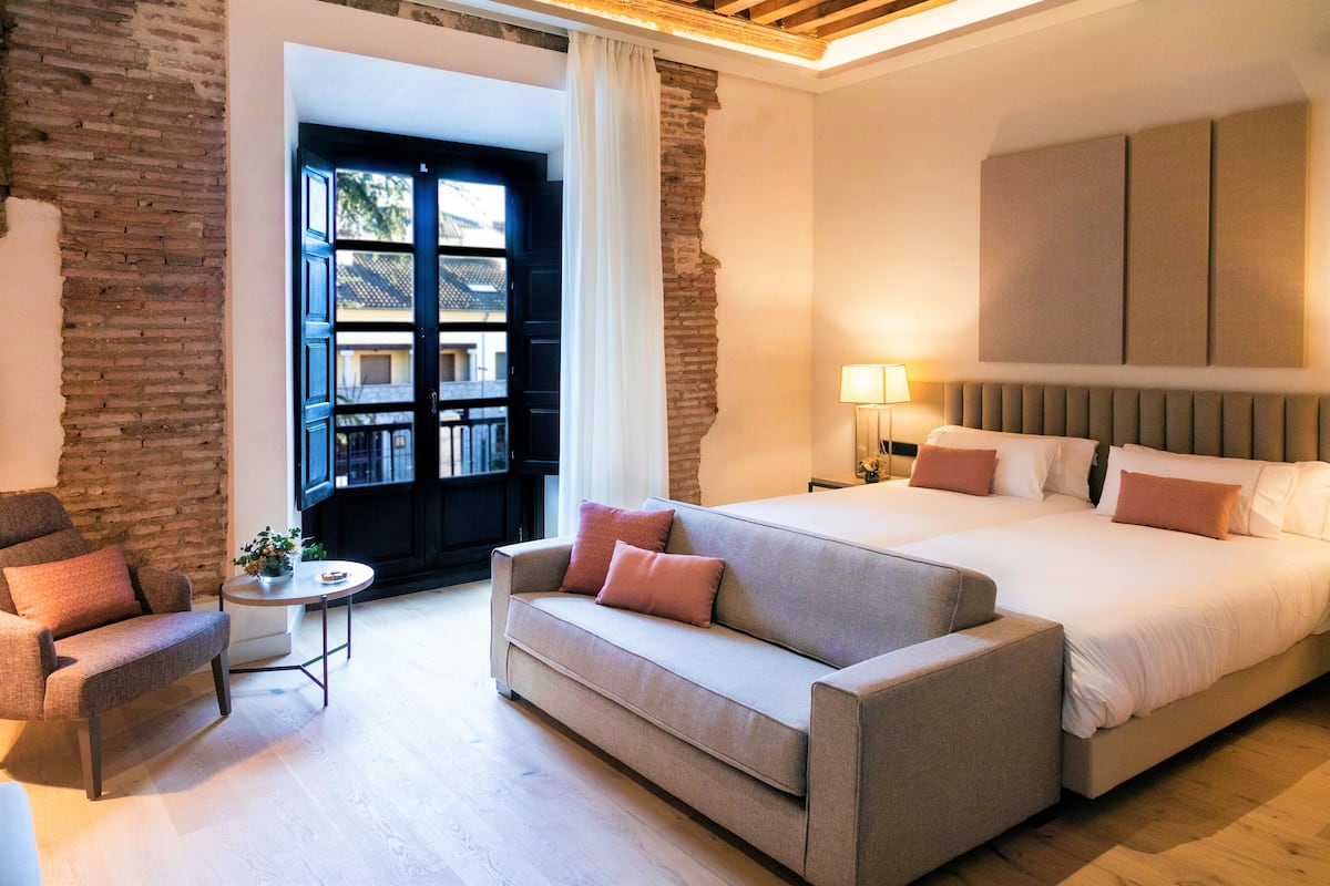 A modern hotel room with a large bed, a sofa, a small table, and a view of the outside.