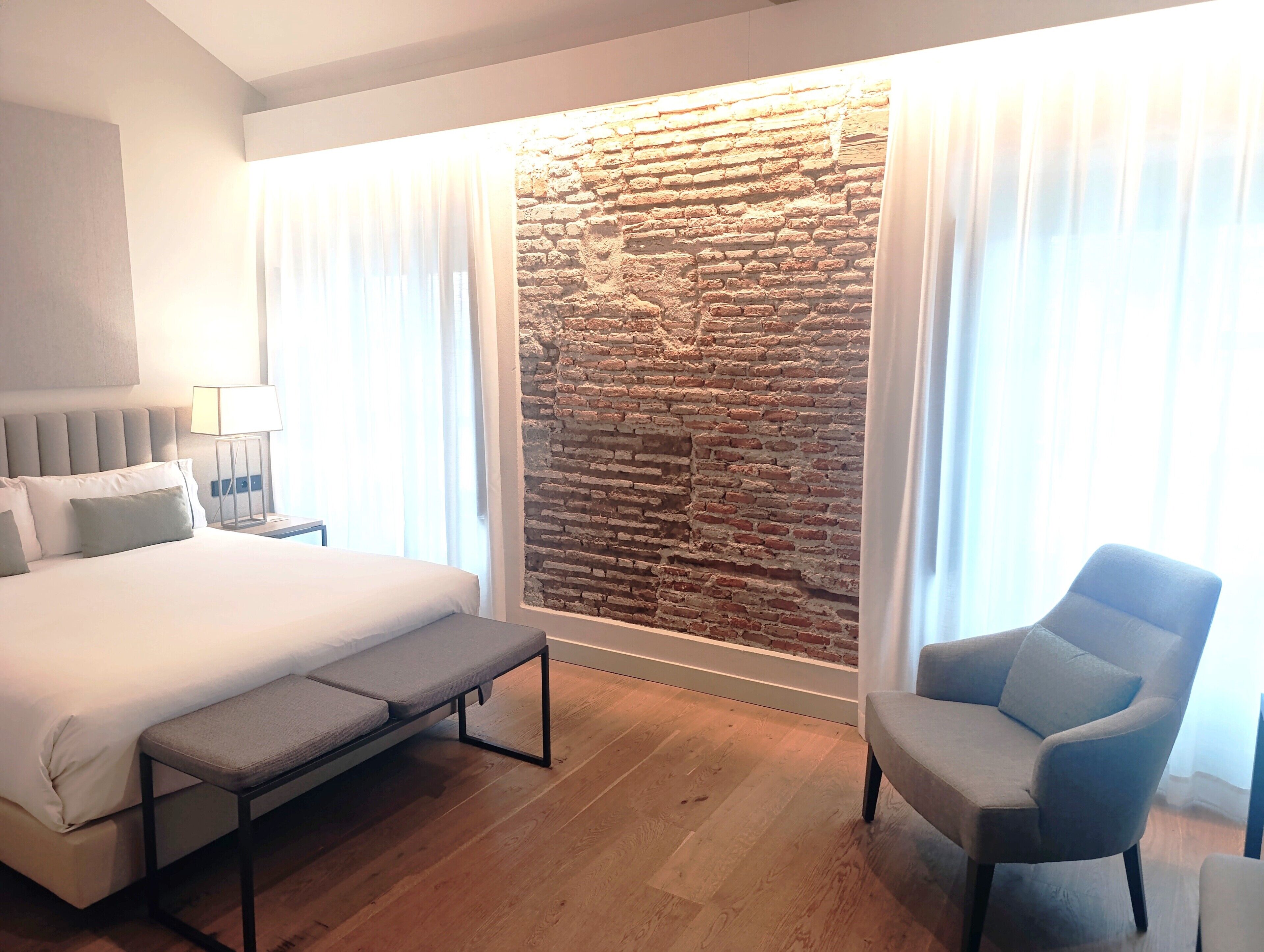 Habitación de hotel con una cama grande, un banco, una silla, una mesita de noche con lámpara y una pared de ladrillo decorativa.