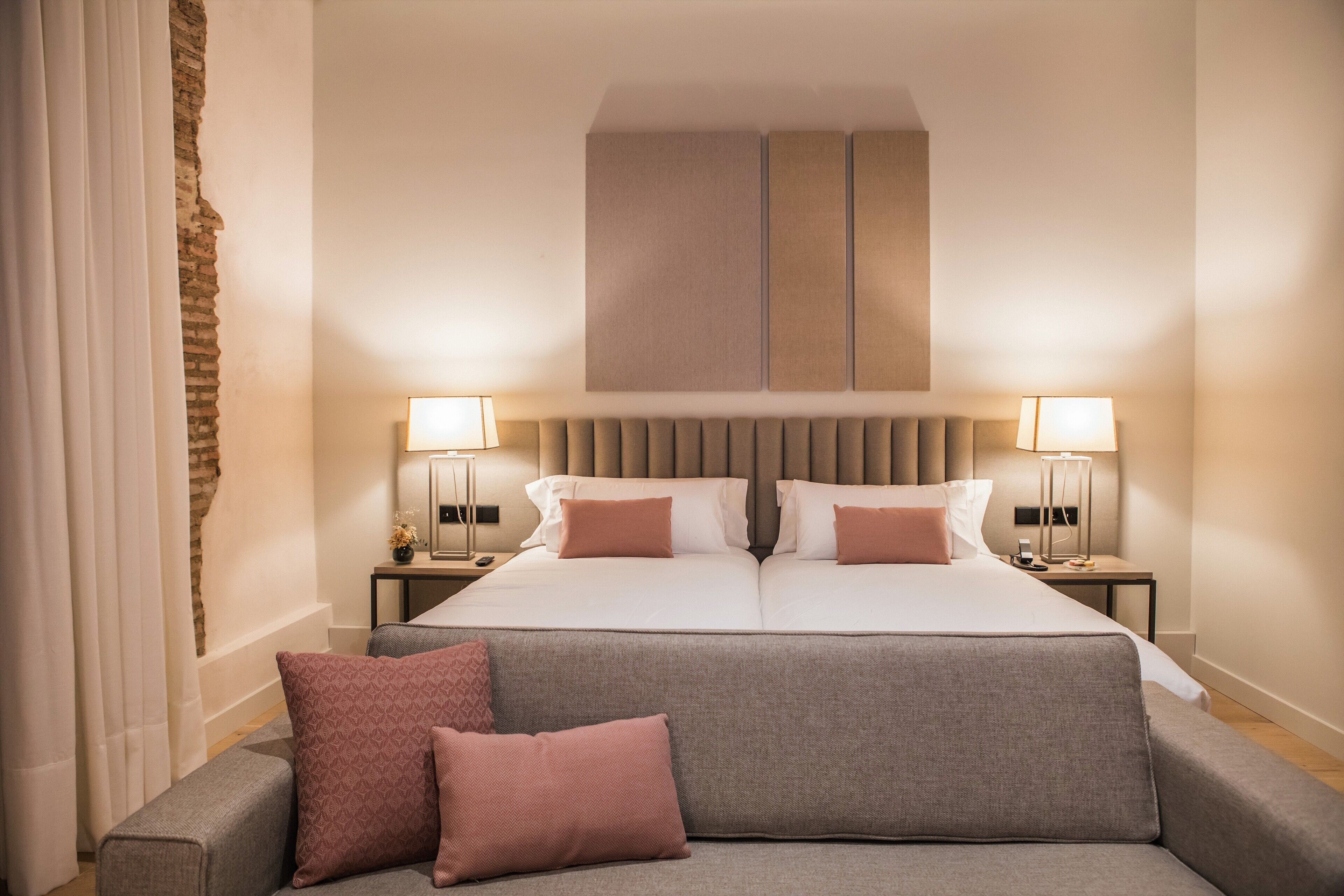 Une chambre d’hôtel avec un grand lit, deux tables de chevet avec des lampes, un canapé gris avec des coussins et un mur en briques apparentes.
