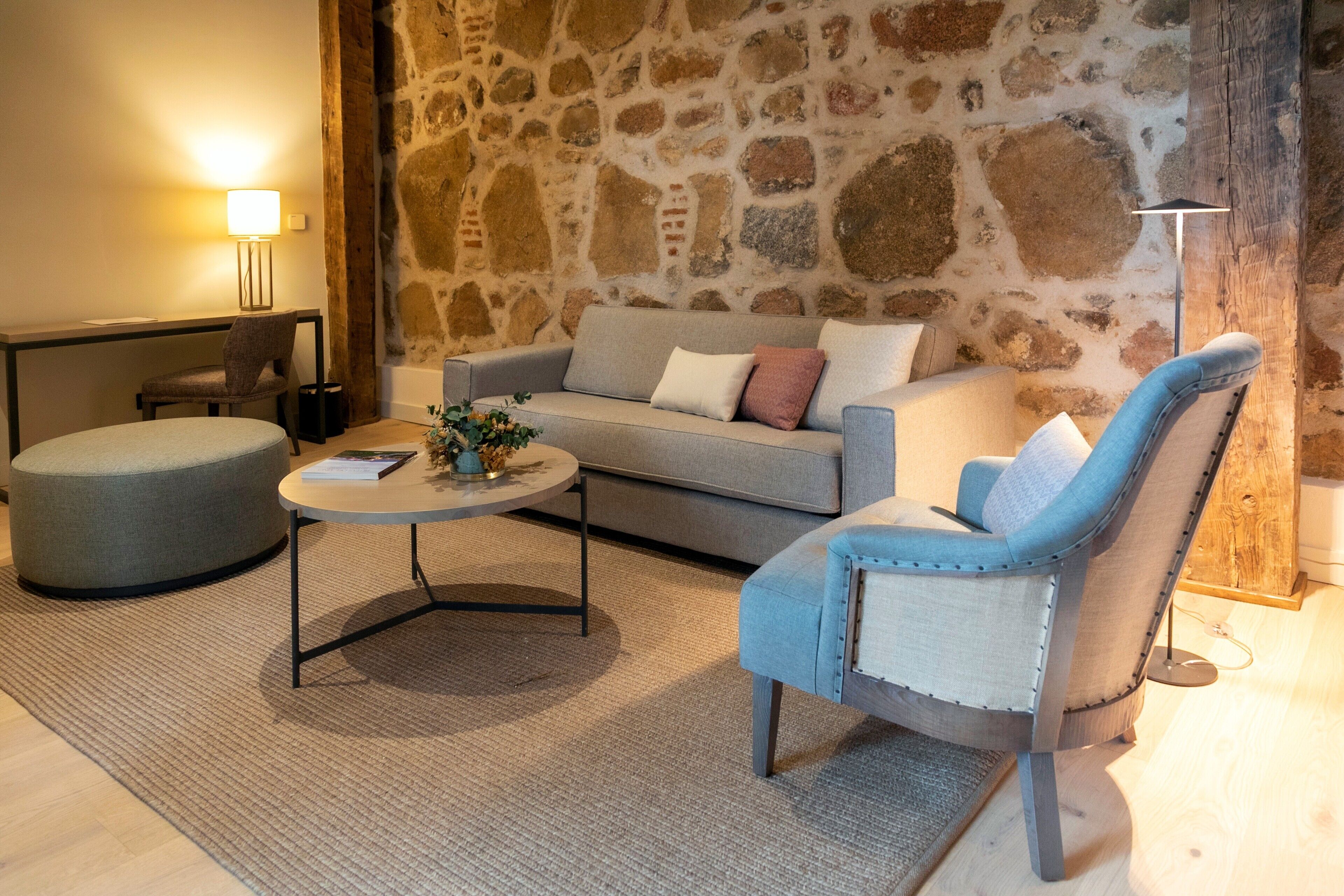 A cozy living room with a stone wall, a grey sofa, a blue armchair, a round coffee table, and a small ottoman.
