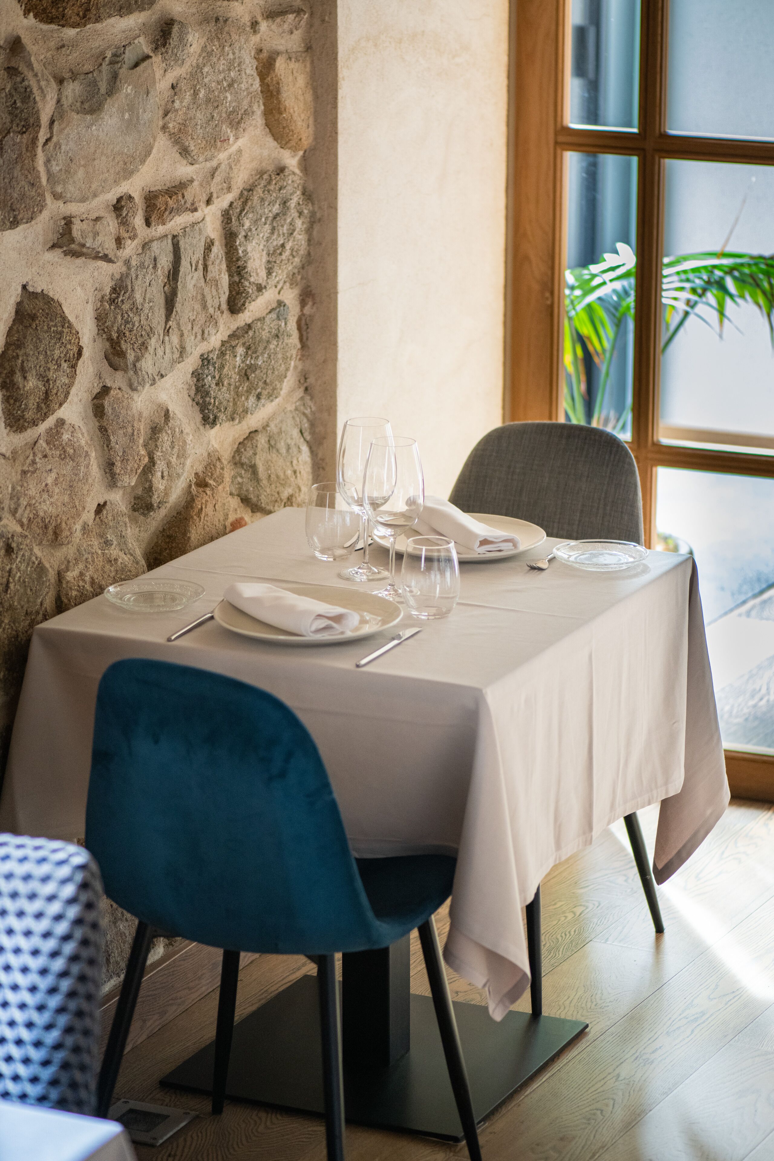 Una mesa de comedor dispuesta con platos, vasos y cubiertos, ubicada frente a una pared de piedra con una ventana al fondo.