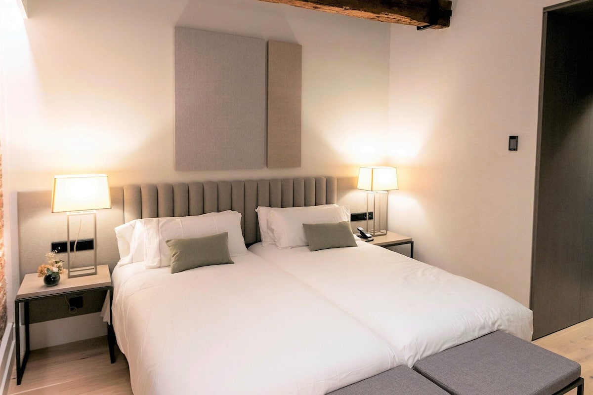 A modern bedroom with a large bed, bedside lamps, a bench, and exposed wooden beams.