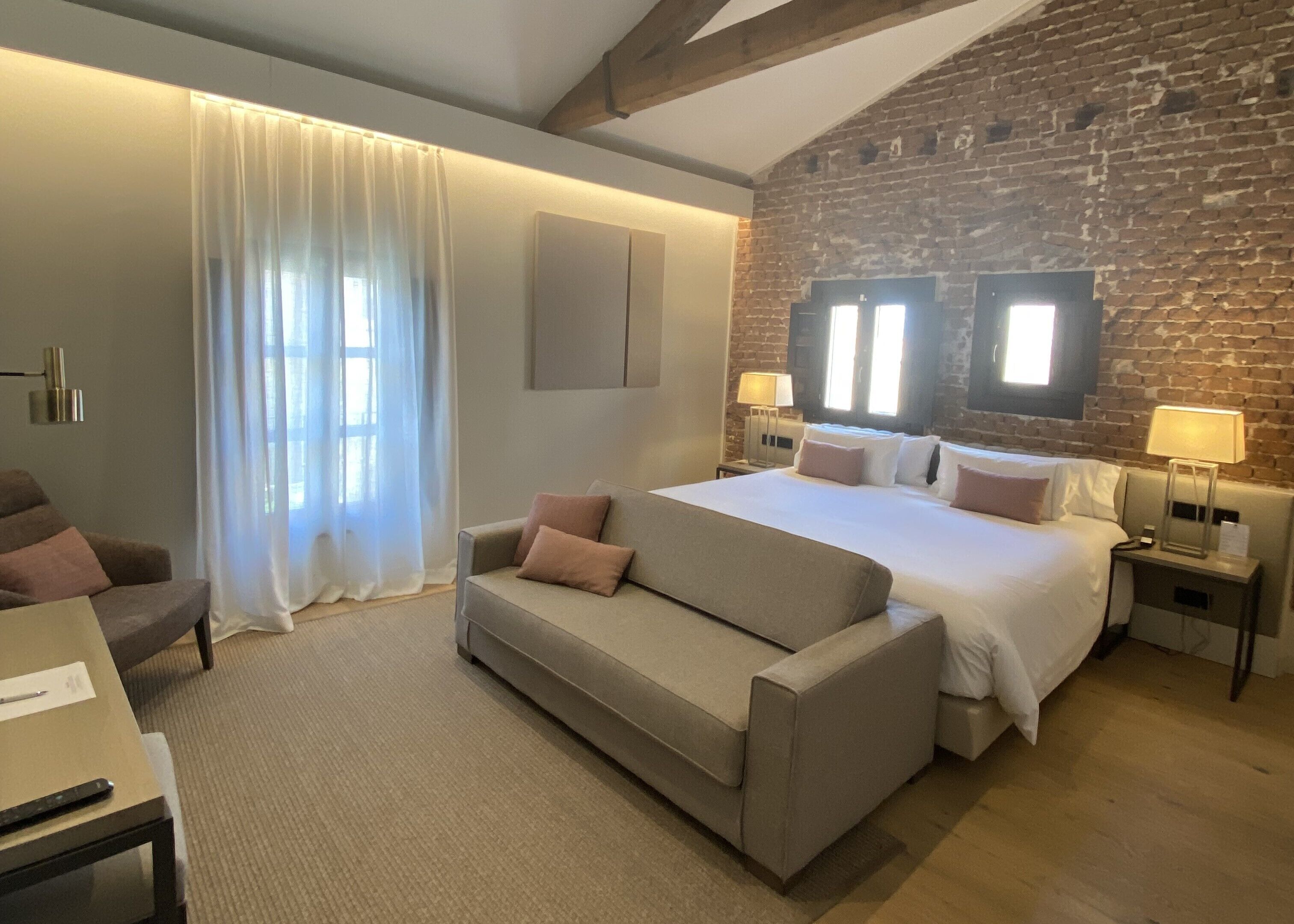 A modern hotel room with a large bed, a sofa, a desk, and a brick wall.