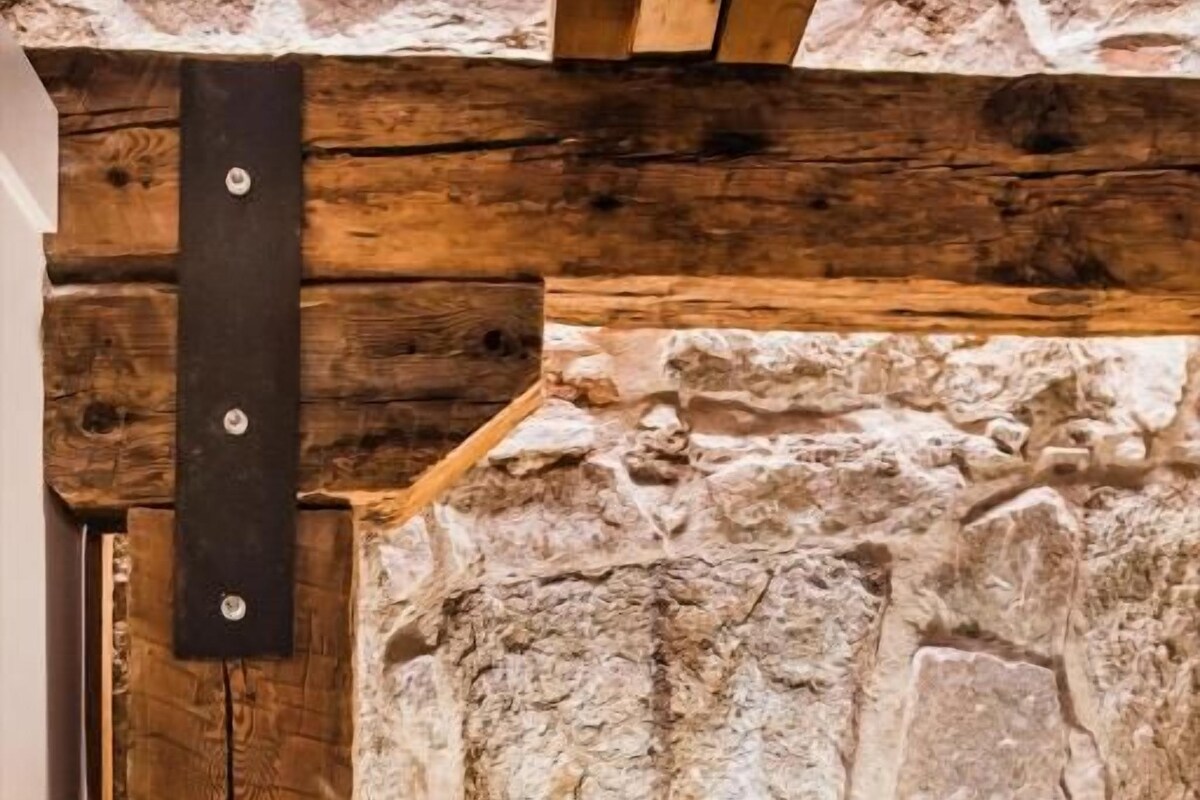 A room with a stone wall, a wooden beam, a bedside lamp, and a white bed.