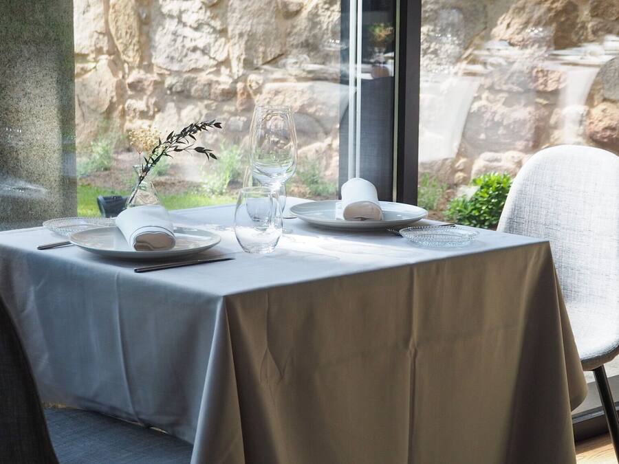 Una mesa de comedor dispuesta con platos, vasos y un florero con una planta, cerca de una ventana que ofrece vistas a un muro de piedra.