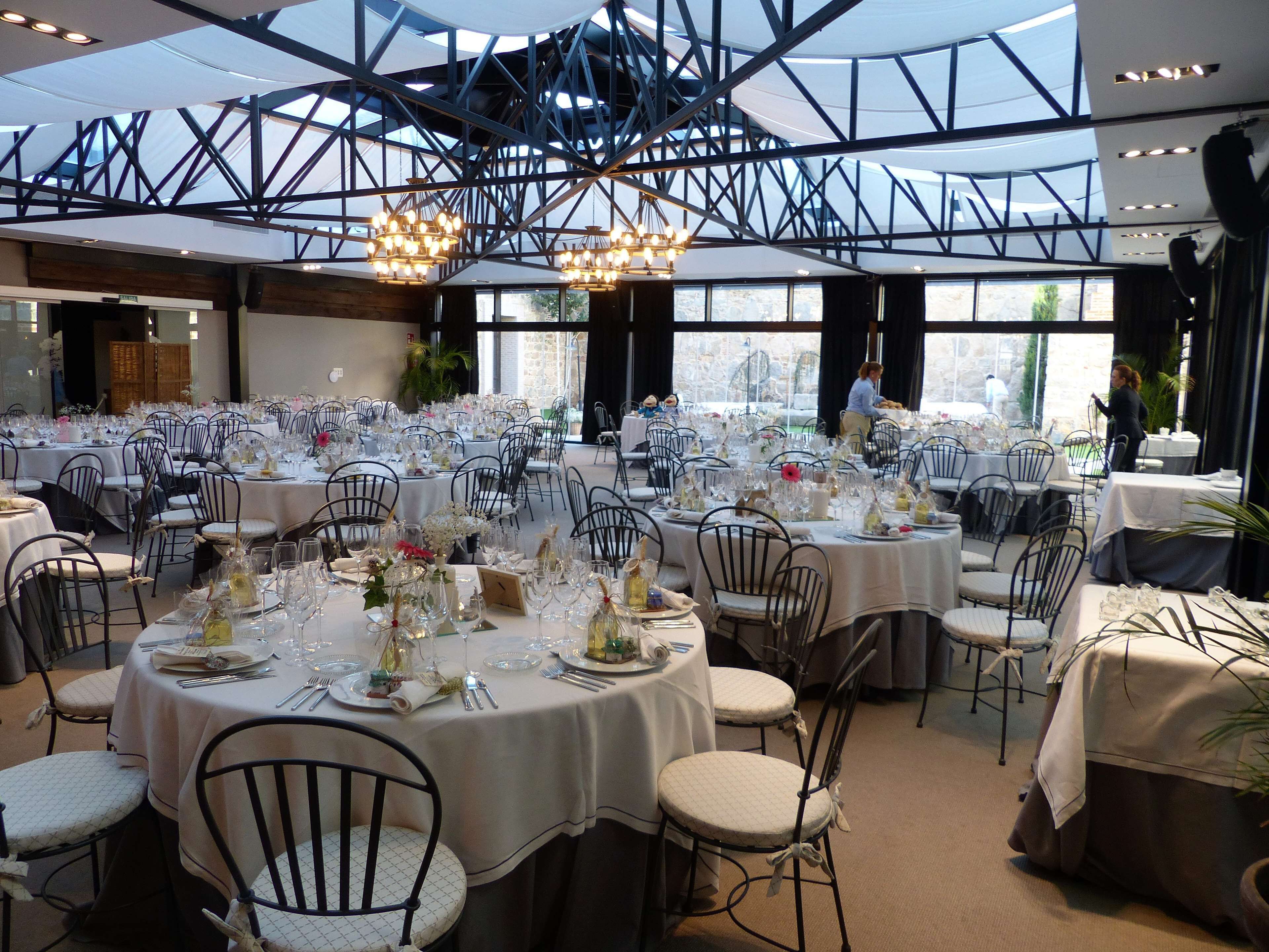 A large indoor dining area with round tables set for a formal event, featuring white tablecloths, glassware, and floral centerpieces.