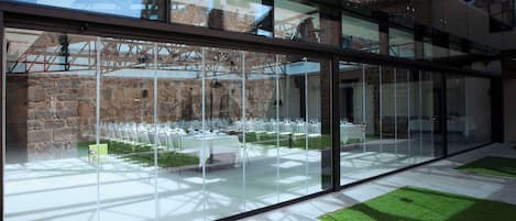 Un área de comedor al aire libre moderna con paredes de vidrio, paredes de piedra y vistas a edificios históricos.