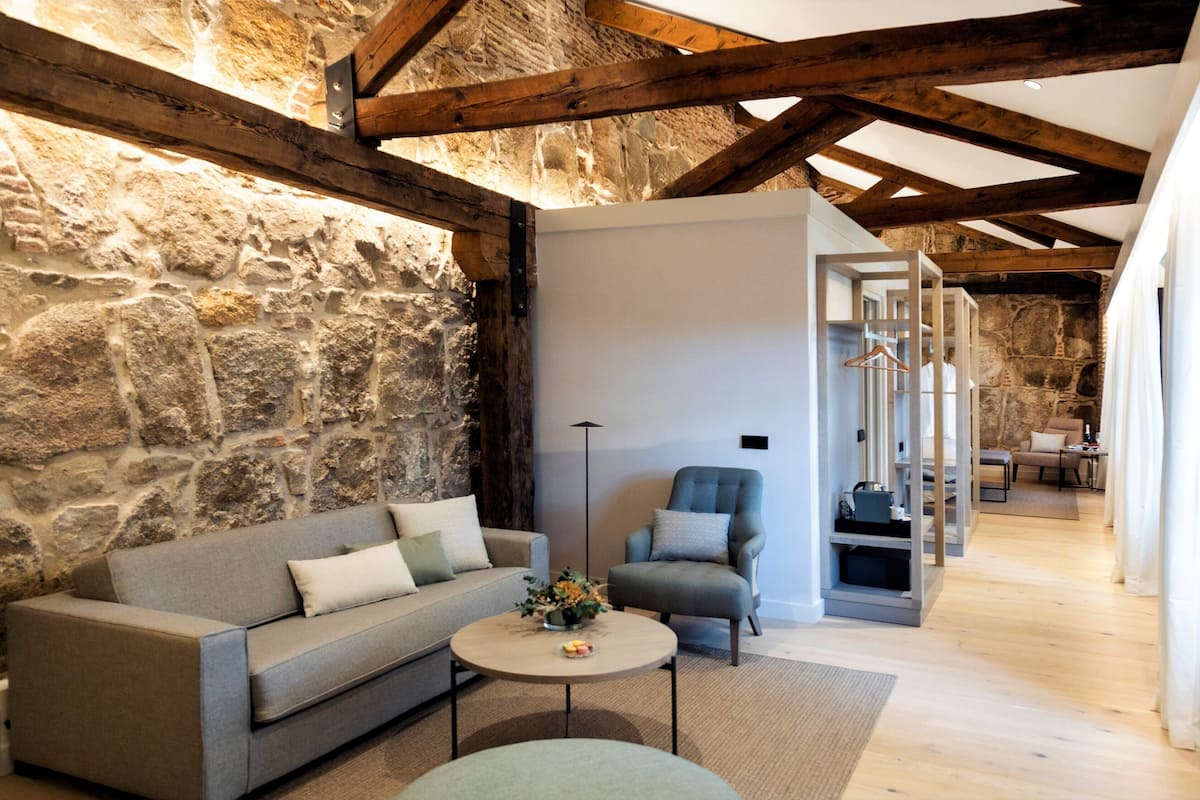 A cozy living room with a stone wall, wooden beams, a grey sofa, a small round table with a vase, and a grey armchair.
