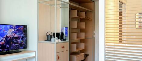 A room with a TV on a white stand, a wooden cabinet with drawers, a shelf with items, and a window with blinds.