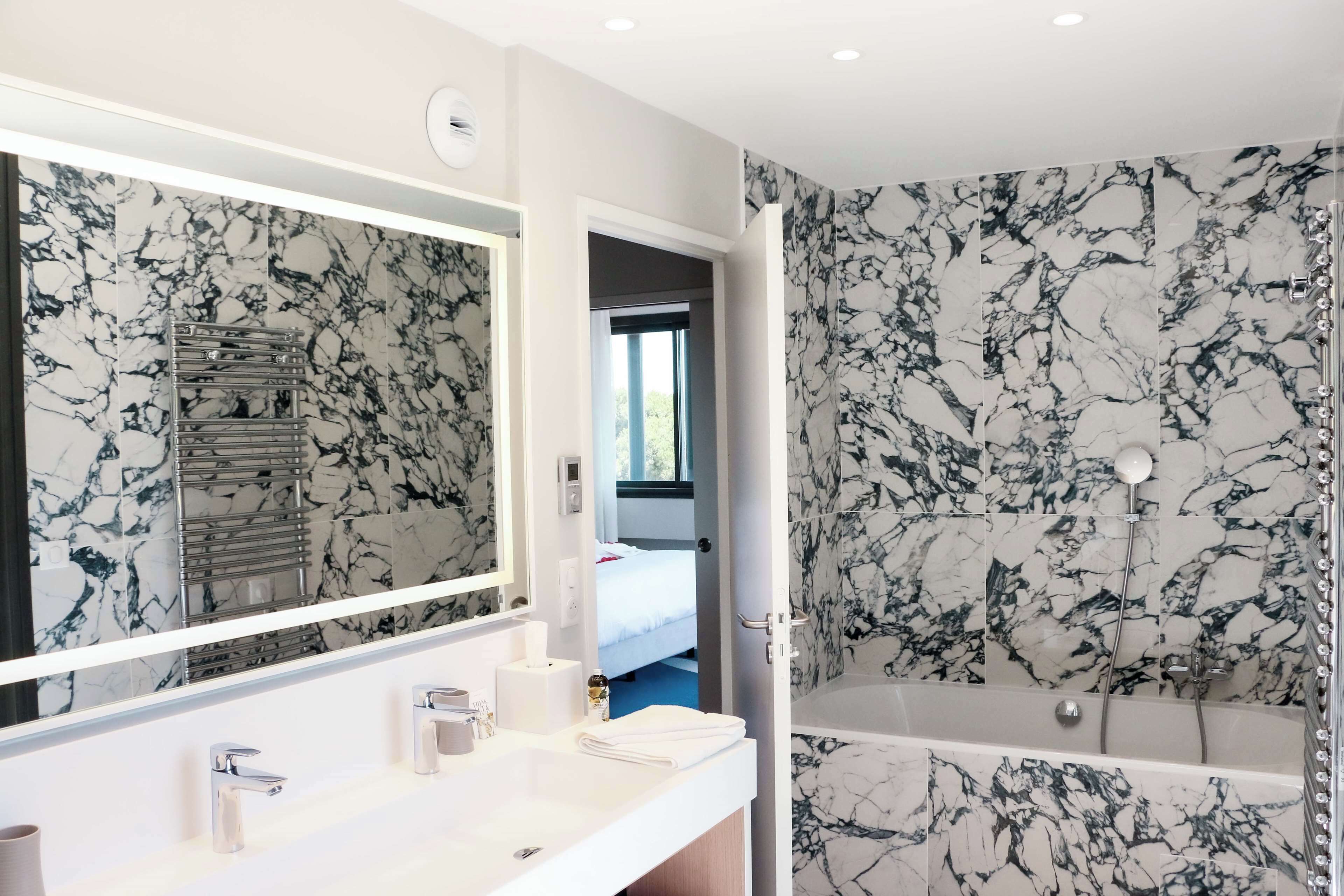 A modern bathroom with a marble backsplash, a large mirror, a white sink with two faucets, and a shower area.
