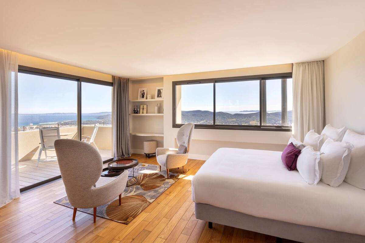 A modern hotel room with a large bed, two armchairs, a small table, and a view of the sea through large windows.