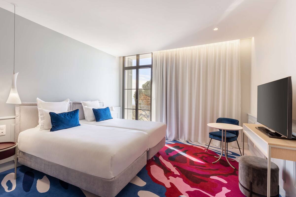 A hotel room with a large bed, a desk with a TV, a chair, and a window with curtains.