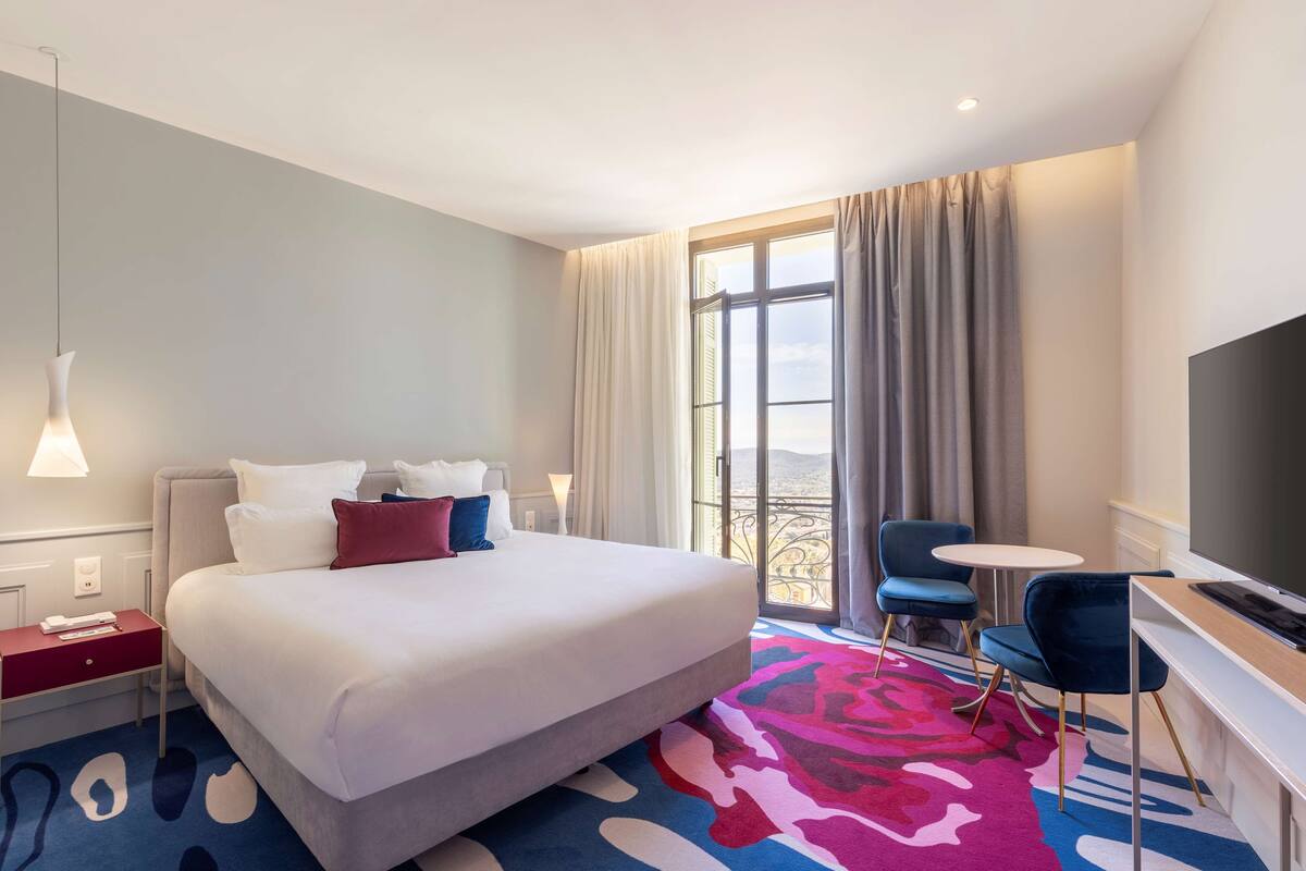 A hotel room with a bed, a TV, a desk, a chair, a window with curtains, and a balcony view.