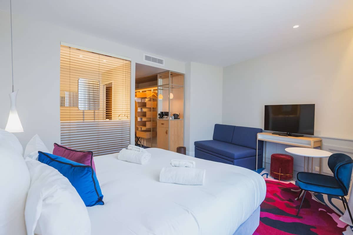 A modern hotel room with a large bed, a TV, a desk, and a seating area.