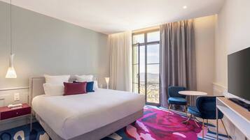 A hotel room with a bed, a TV, a desk, a chair, and a balcony with curtains.
