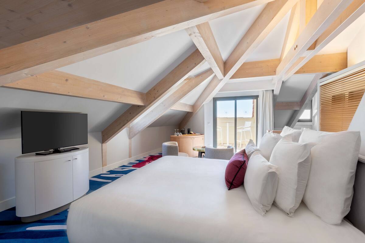 A modern hotel room with a large bed, a TV, a desk, and a window with blinds.