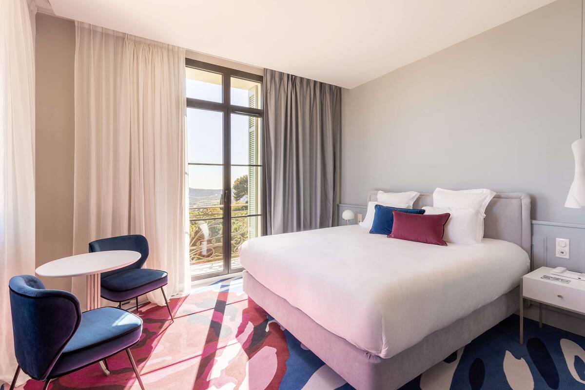 A hotel room with a large bed, a small table, two chairs, and a view of the outdoors through a large window.