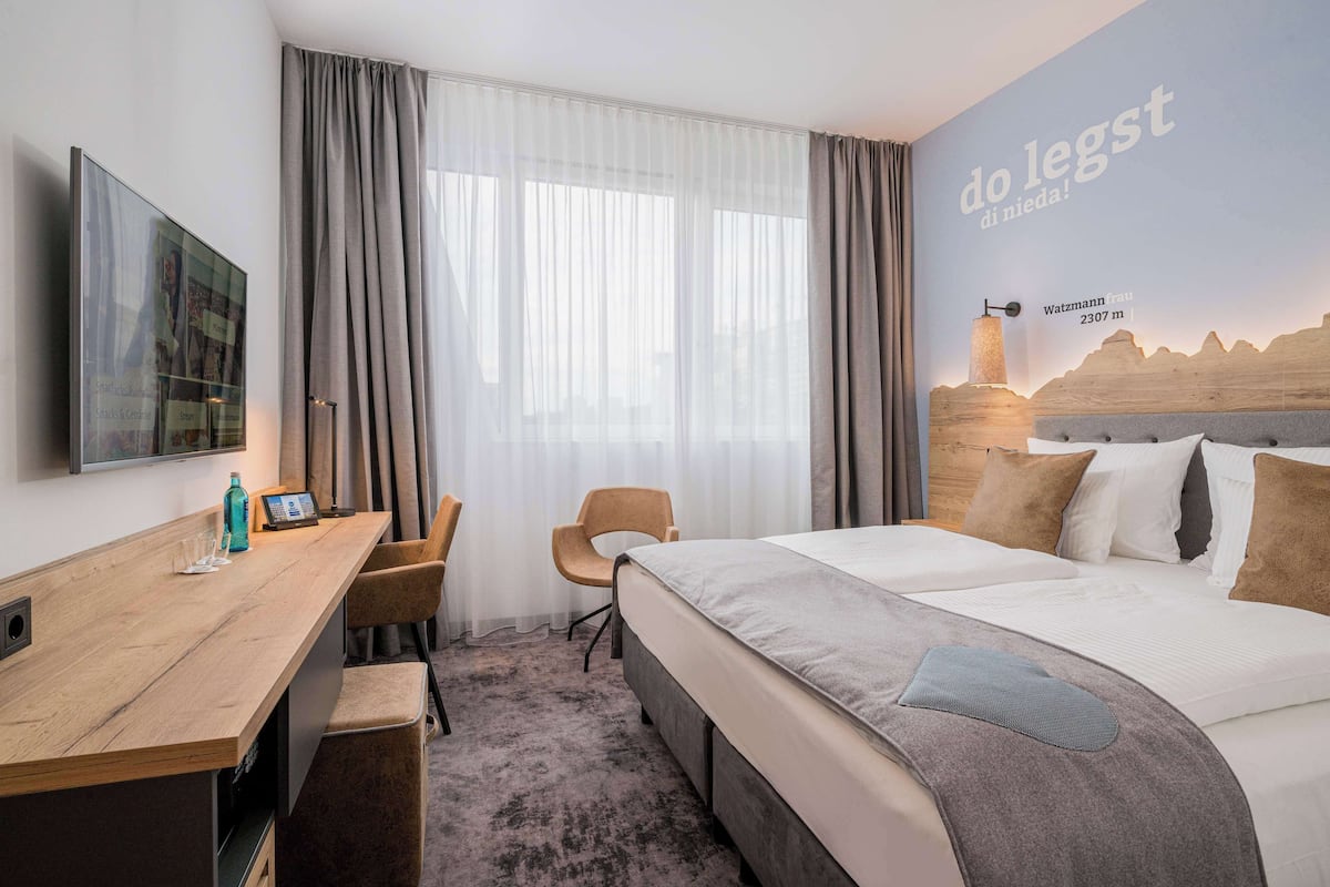 Hotelkamer met een bed, bureau, stoel, televisie en een muur met een berg en tekst.