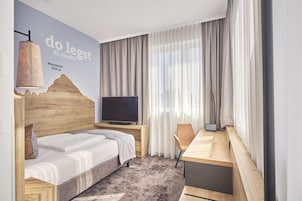 Et hotelværelse med seng, skrivebord, fjernsyn og et stort vindue med gardiner.