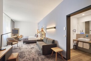 A modern hotel room with a bed, sofa, desk, and a bathroom with a sink and mirror.
