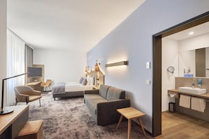 Habitación de hotel moderna con cama, sofá, escritorio y baño con lavamanos y espejo.