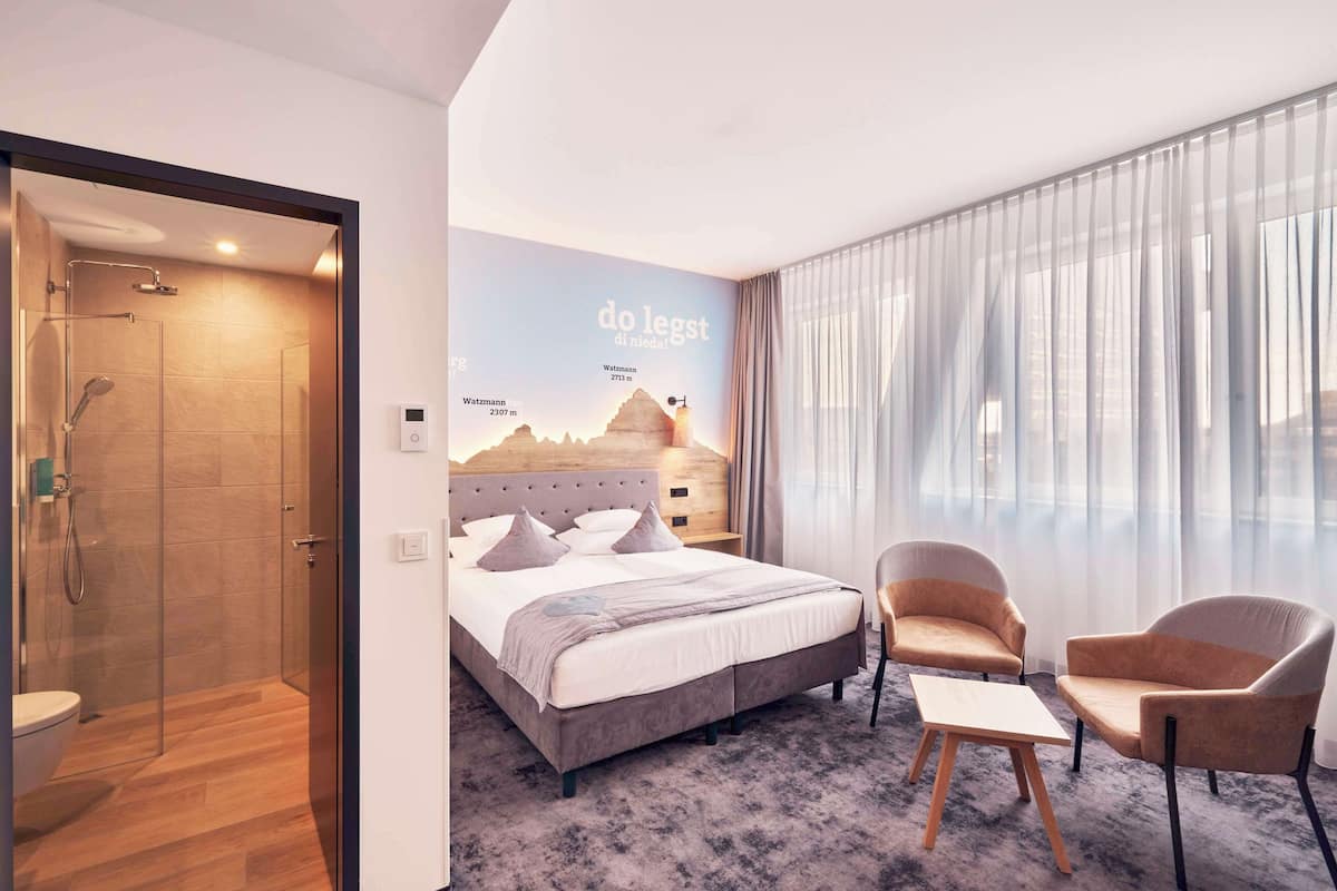 Een moderne hotelkamer met een groot bed, twee fauteuils, een tafeltje, een douche en een muurschildering van een berg.
