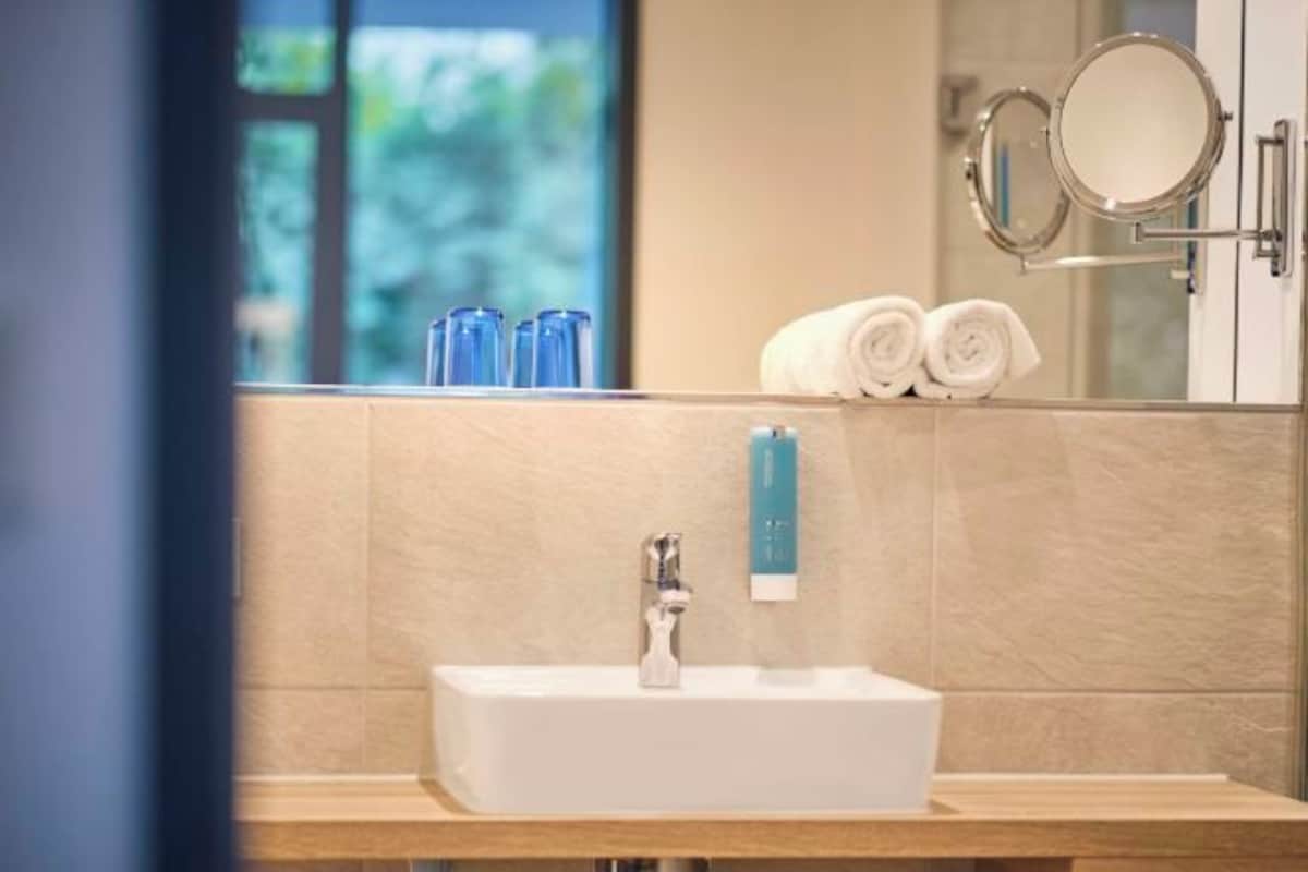 Une salle de bain avec un lavabo, deux miroirs et une fenêtre donnant sur un paysage verdoyant.