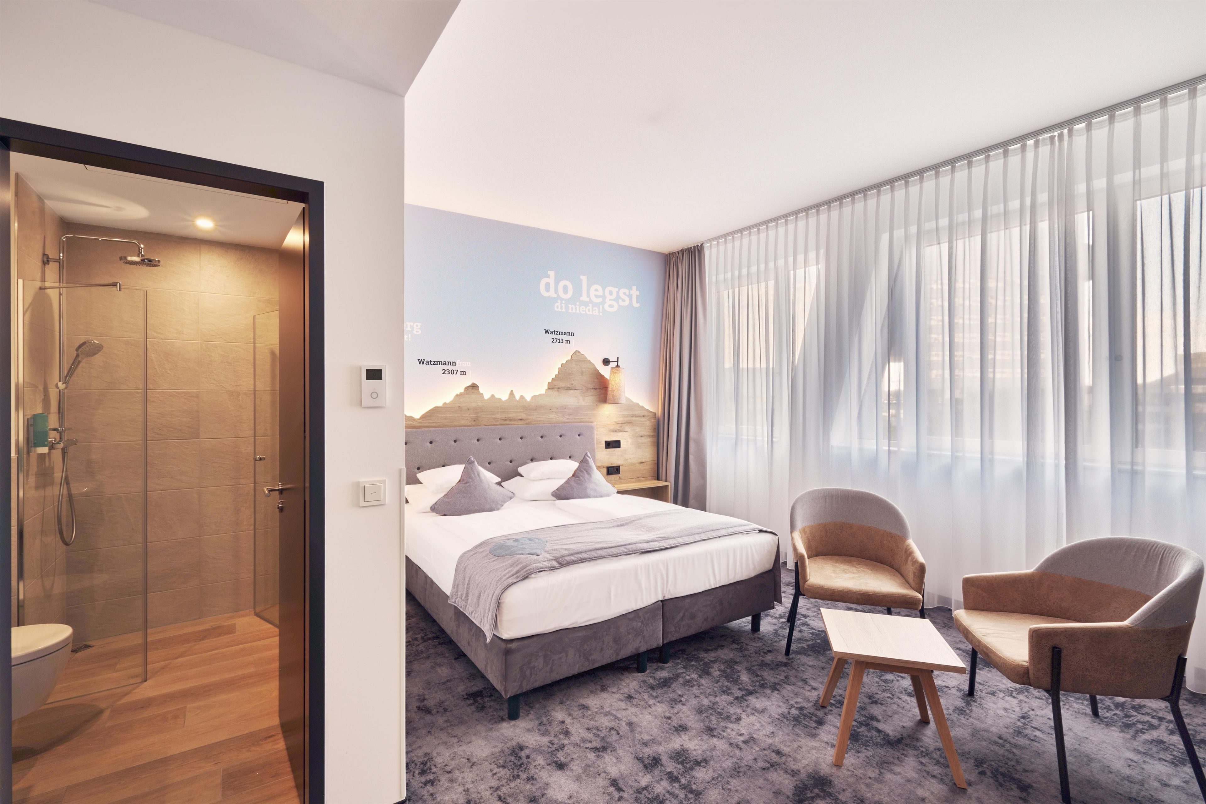 Ein Hotelzimmer mit einem Bett, zwei Sesseln, einem kleinen Tisch, einer Duschkabine und einem Wandbild von Bergen.