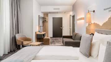 Ein Hotelzimmer mit einem Bett, zwei Sesseln, einem kleinen Tisch, einem Schreibtisch, einem Spiegel, einer Lampe und einem Wandhandtuchhalter.
