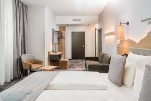 Une chambre d’hôtel comprenant un lit, deux fauteuils, une petite table, un bureau, un miroir, une lampe et un porte-serviettes fixé au mur.