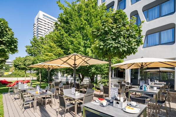 An outdoor dining area with tables and chairs, umbrellas, and a view of a building with blue windows. - Best Western Hotel Arabellapark München (Munich)