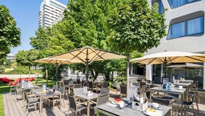 An outdoor dining area with tables and chairs, umbrellas, and greenery.
