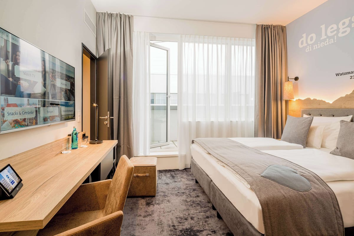 Hotelkamer met een bed, een bureau met een tablet, een stoel, een raam met gordijnen en een muur met een poster.