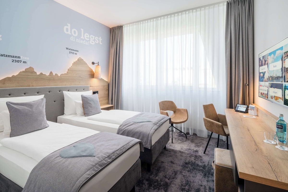 Een hotelkamer met twee bedden, een houten hoofdbord, een bureau met stoel en een groot raam met dunne gordijnen.