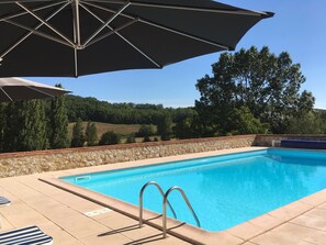 Una piscina con muro de piedra y solárium con baldosas, rodeada de árboles y cielo despejado.