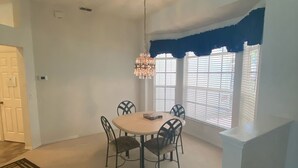 A dining area with a round table, six chairs, a chandelier, and a window with blinds.