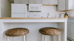 Eine Küche mit weißen Schränken, einem weißen Kühlschrank, einer Mikrowelle und einer Holz Arbeitsplatte. Zwei gewebte Hocker stehen an der Theke.