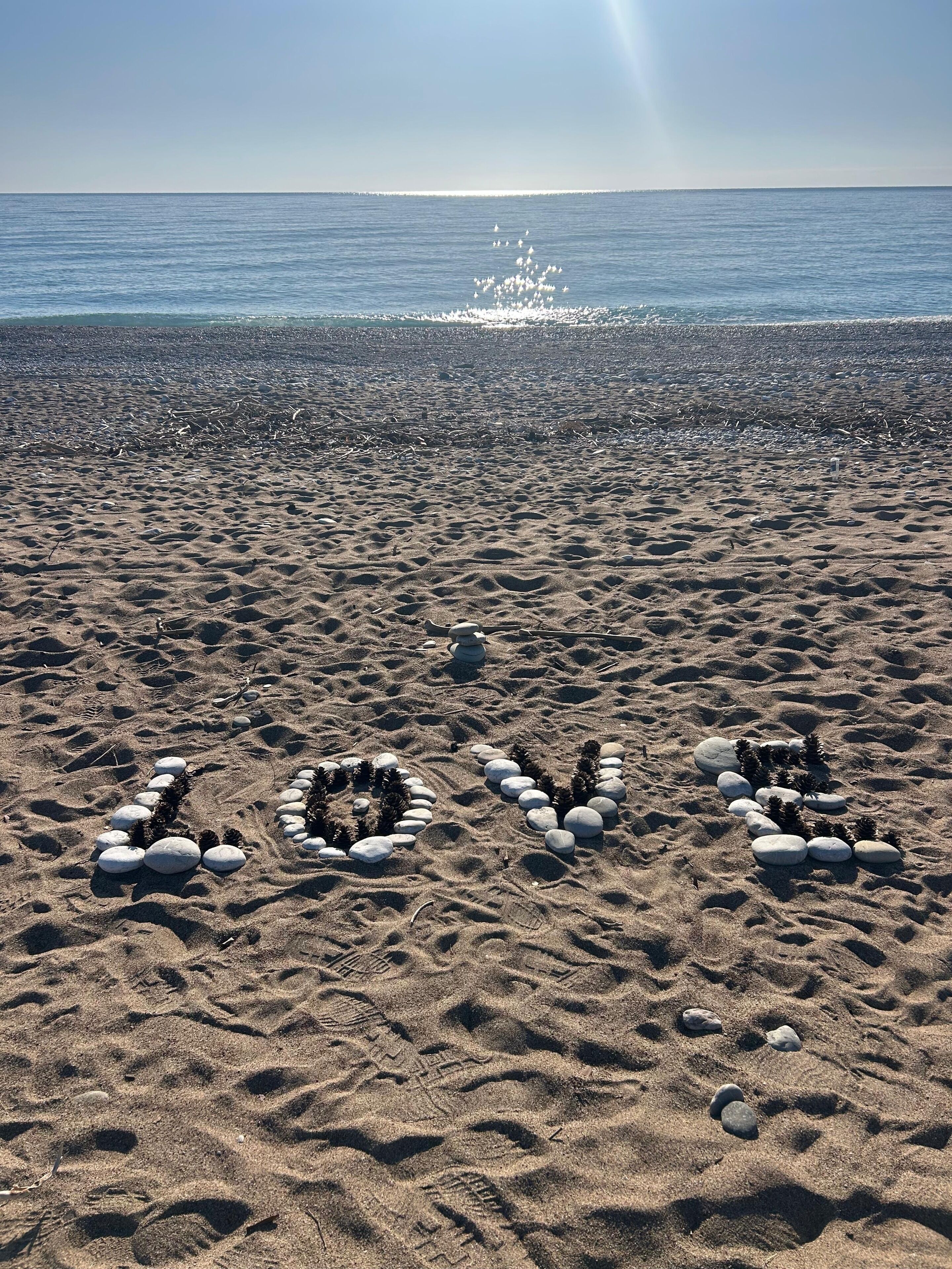Une plage de sable où des pierres sont disposées de manière à former le mot « AMOUR », et où l’on peut voir des empreintes de pas dans le sable.