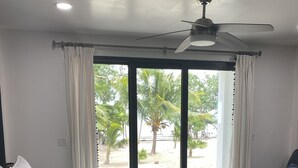 A bedroom with a bed, a ceiling fan, and a view of palm trees and a beach through large windows. - Shedrock Shangrila, Beachfront Ocean Breeze Suite (Hopkins)