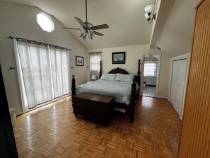 Relax in this serene bedroom with a large bed and ceiling fan, surrounded by natural light and a beautiful wooden floor.