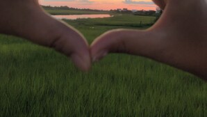Handen vormen een hart voor een zonsondergang boven een grasveld.