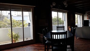 A dining area with a wooden table and chairs, large windows with a view of buildings and trees, and a television on a stand. - Habitación Privada // Casa (DRN)