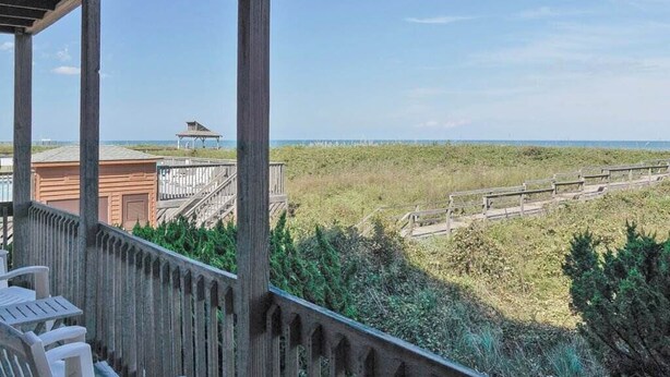 A wooden deck with chairs, overlooking a beach with a wooden fence and a distant structure. - Charming first floor condo in Duck! (Duck)