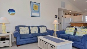 A living room with two blue sofas, a white coffee table, and a kitchen in the background. - Enjoy partial ocean views from this tastefully decorated home in Nags Head. (Nags Head)