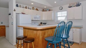 A compact kitchen with a central island, two bar stools, and a window with blinds. - Enjoy partial ocean views from this tastefully decorated home in Nags Head. (Nags Head)