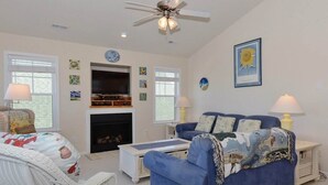 A living room with a blue sofa, white wicker chair, a fireplace, and a ceiling fan. - Enjoy partial ocean views from this tastefully decorated home in Nags Head. (Nags Head)