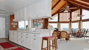 Uma cozinha espaçosa com armários brancos, uma ilha central com banquetas e vista para o oceano através de grandes janelas.