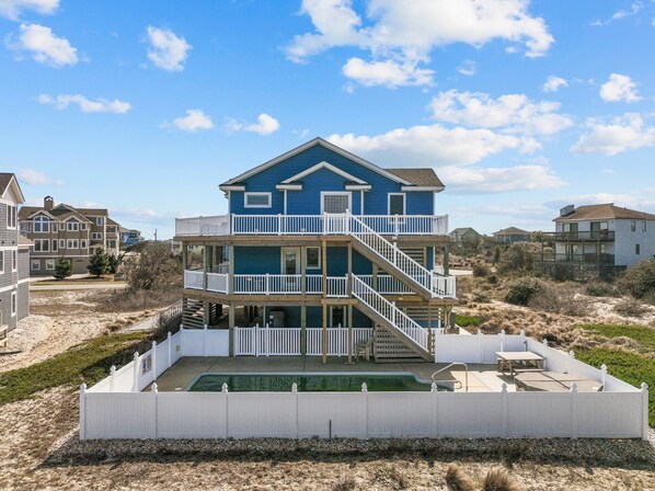 Una casa frente a la playa de dos pisos con piscina, barandillas blancas y una playa arenosa al fondo.