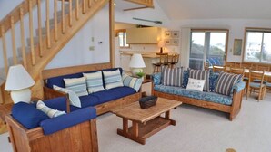 Una sala de estar acogedora con una escalera de madera, dos sofás con cojines a rayas, una mesa de centro de madera y un comedor con sillas y mesa de madera.